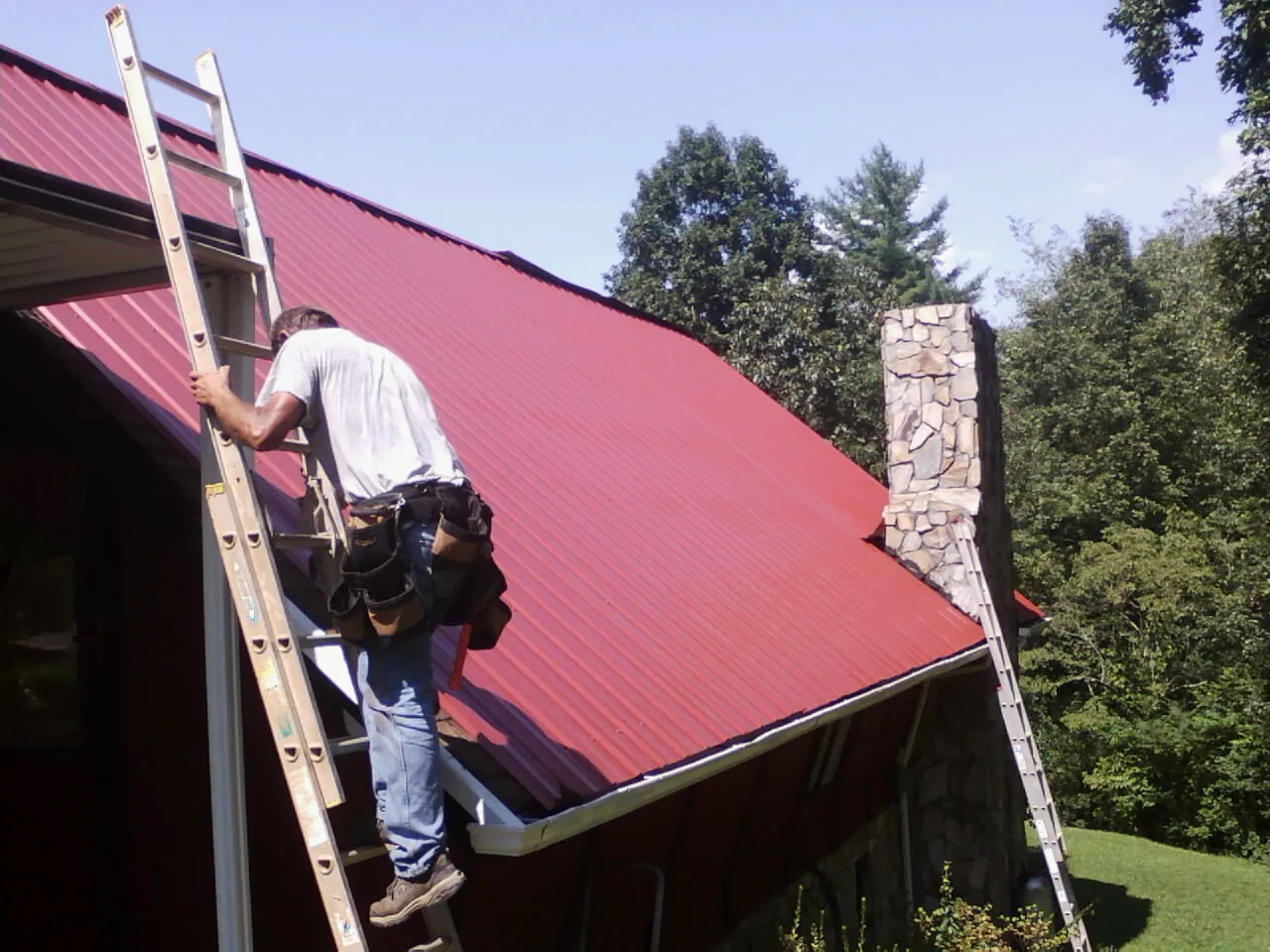 Roofer on a ladder beside a red metal roof and stone chimney — Soffit & Fascia Repair by Roofing Brothers in Wallsburg